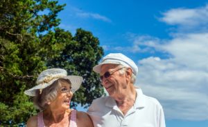 Two older adults smiling at each other. They both wear hats and stand outside. A bright blue sky with a couple clouds and a lush green tree are behind them.