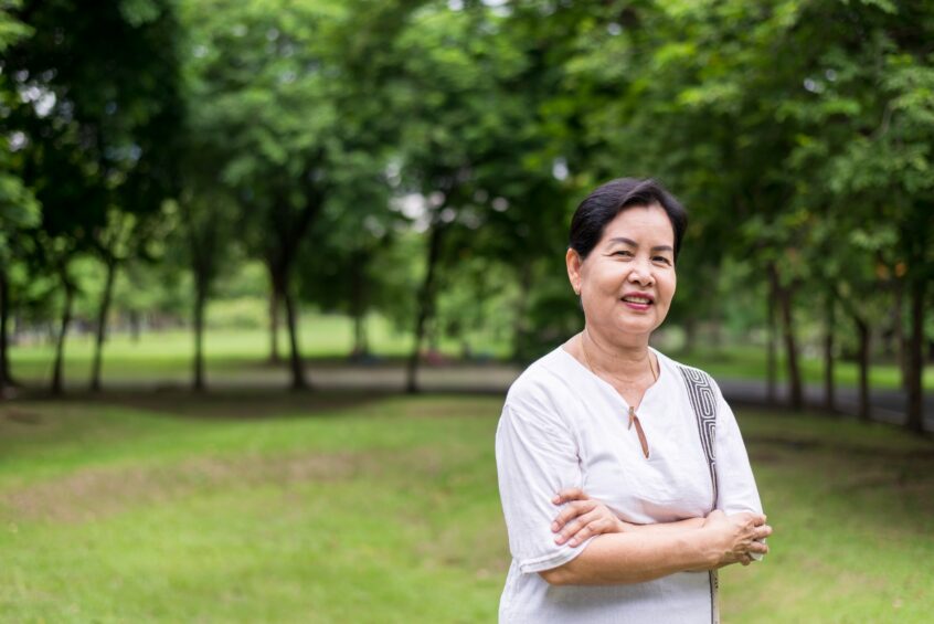 An elderly woman smiling with her arms crossed in front of her. She stands in a wooded park.