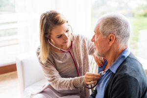 Home health aide listening through stethoscope to senior man’s heart.